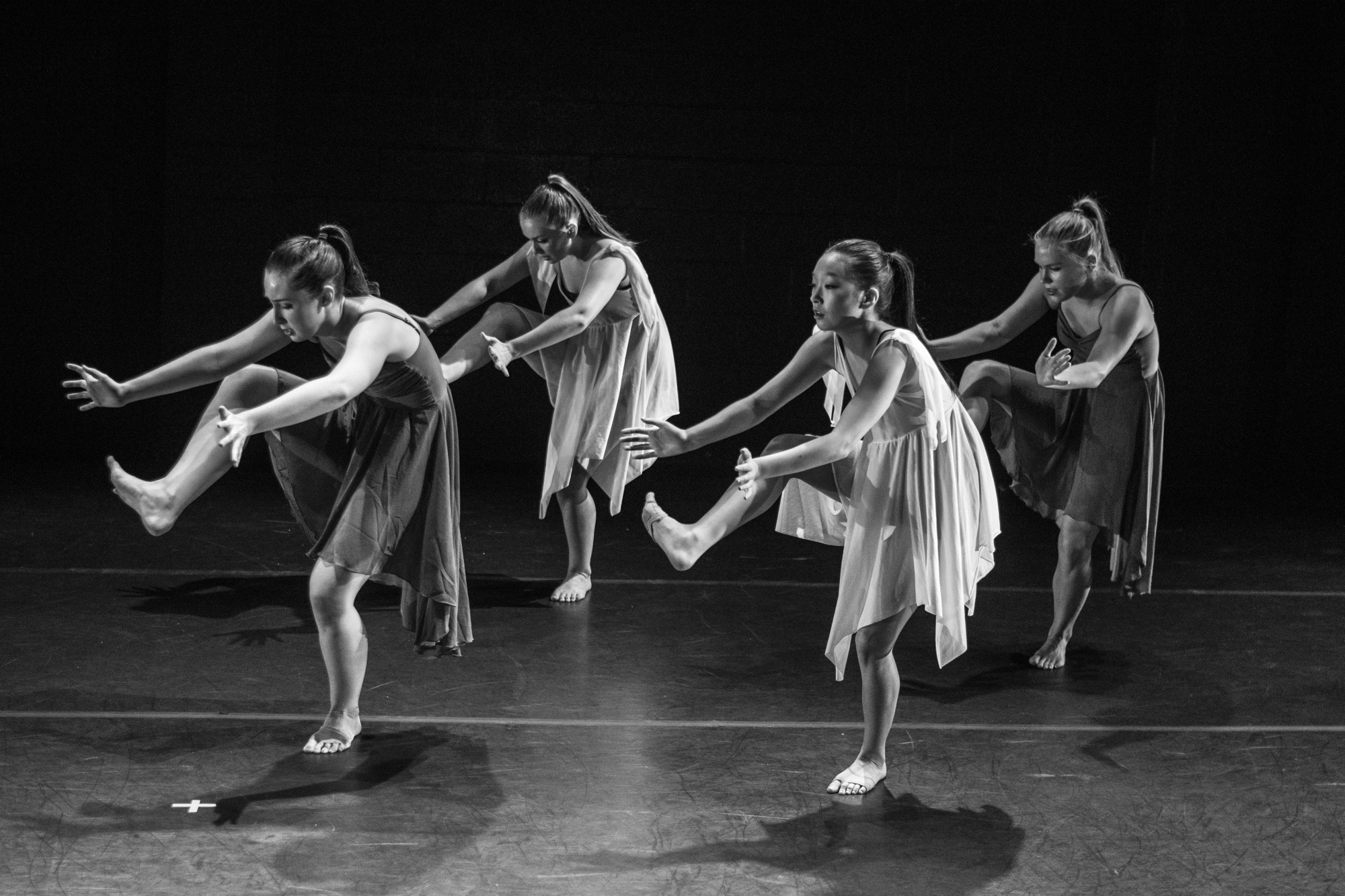 Four dancers in flowing dresses mid-step on a dark stage, black and white